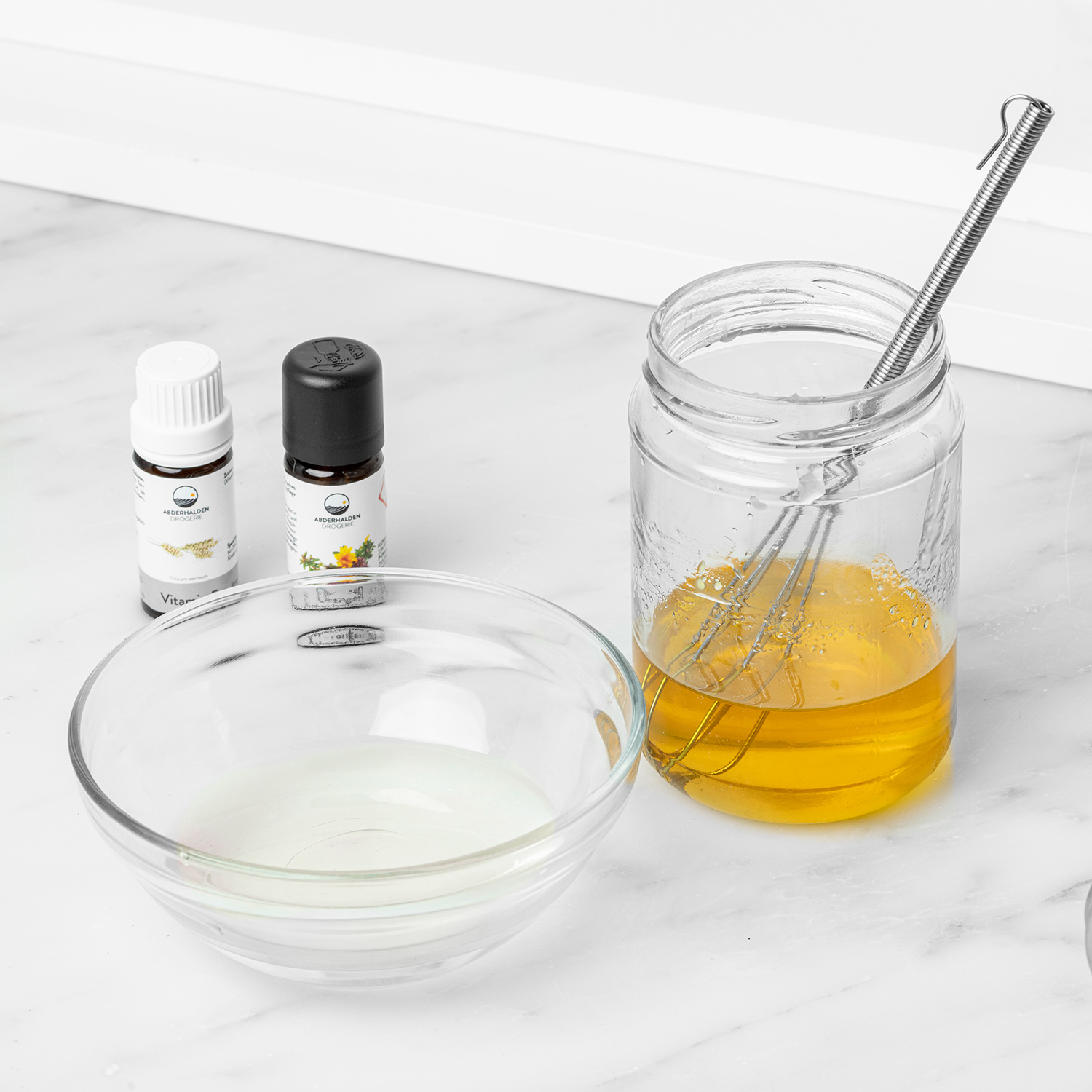 Orangeblütenwasser in Schale neben geschmolzener Fettphase, Vitamin E und Ätherisches Öl dahinter