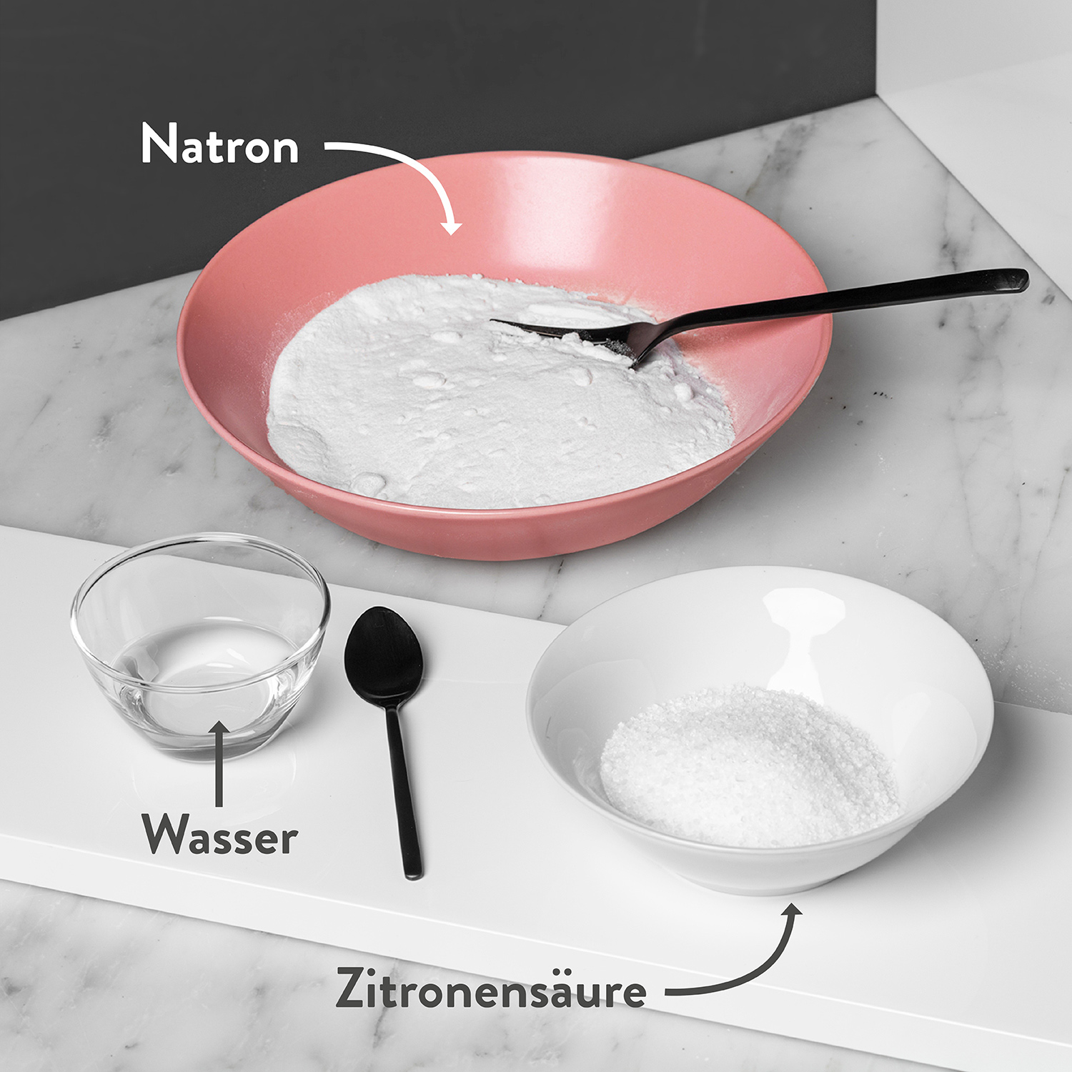Natron in roter Schale, Zitronensäure in weisser Schale und Wasser in Glasschale auf Tisch